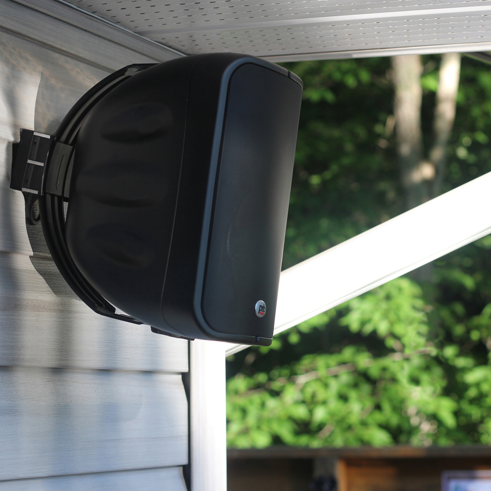 Close-up of a black CS1000 speaker mounted on an exterior wall over vinyl siding, under the soffit