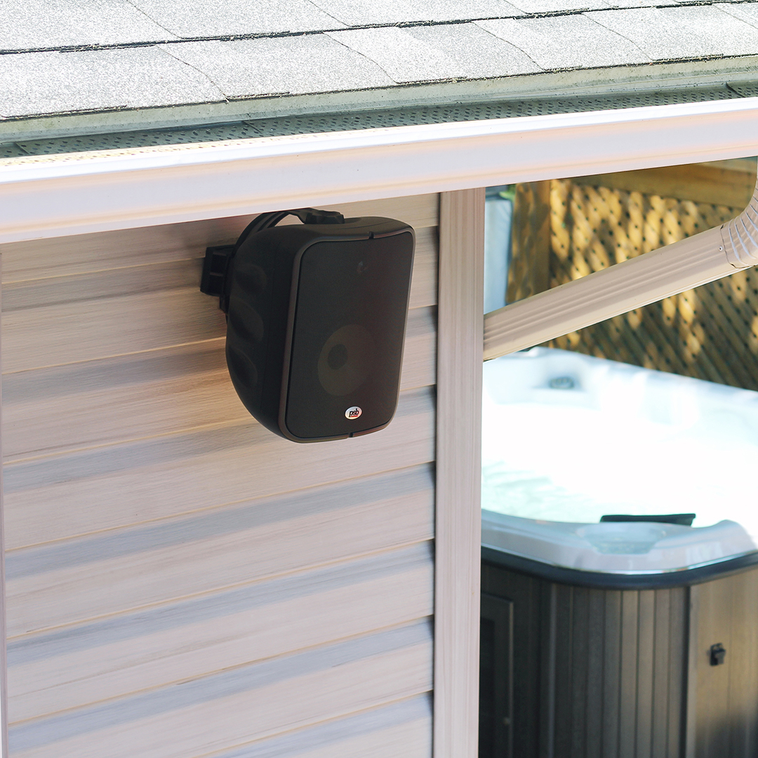 Three-quarter view of a black CS1000 speaker mounted on the outside wall of a home, under the soffit