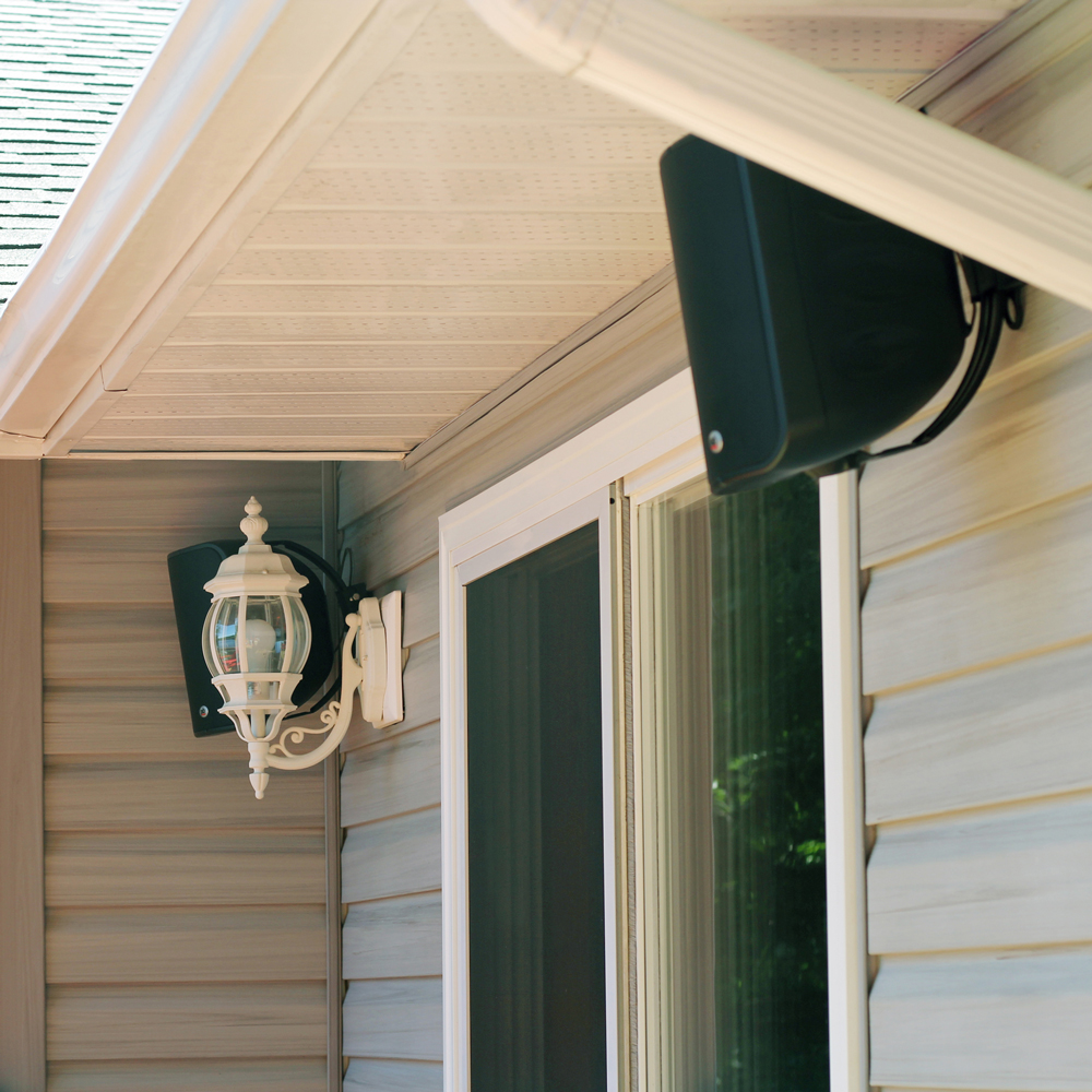 A pair of black CS1000 speakers mounted on the outside of a home, on either side of a sliding glass door