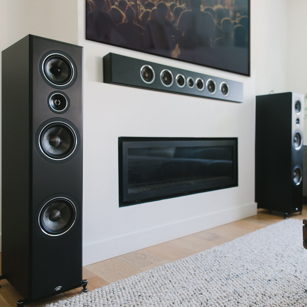 A black PSB PWM3 speaker mounted horizontally on a wall below a large TV, with two other tower speakers standing on the floor